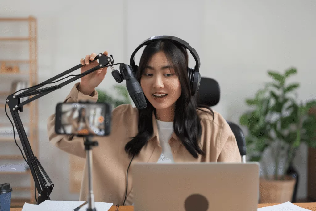 A young asian woman with headphones and a microphone in front of a laptop.