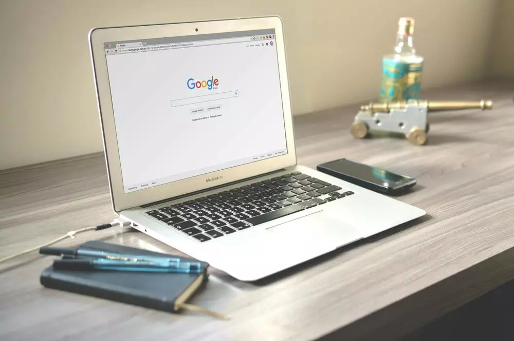 Laptop on a wooden desk with Google search open, alongside a smartphone, notebook, and decorative items.