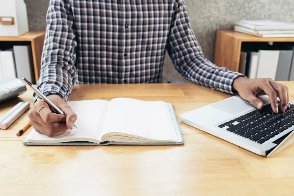 Person writing in a notebook while using a laptop at a desk