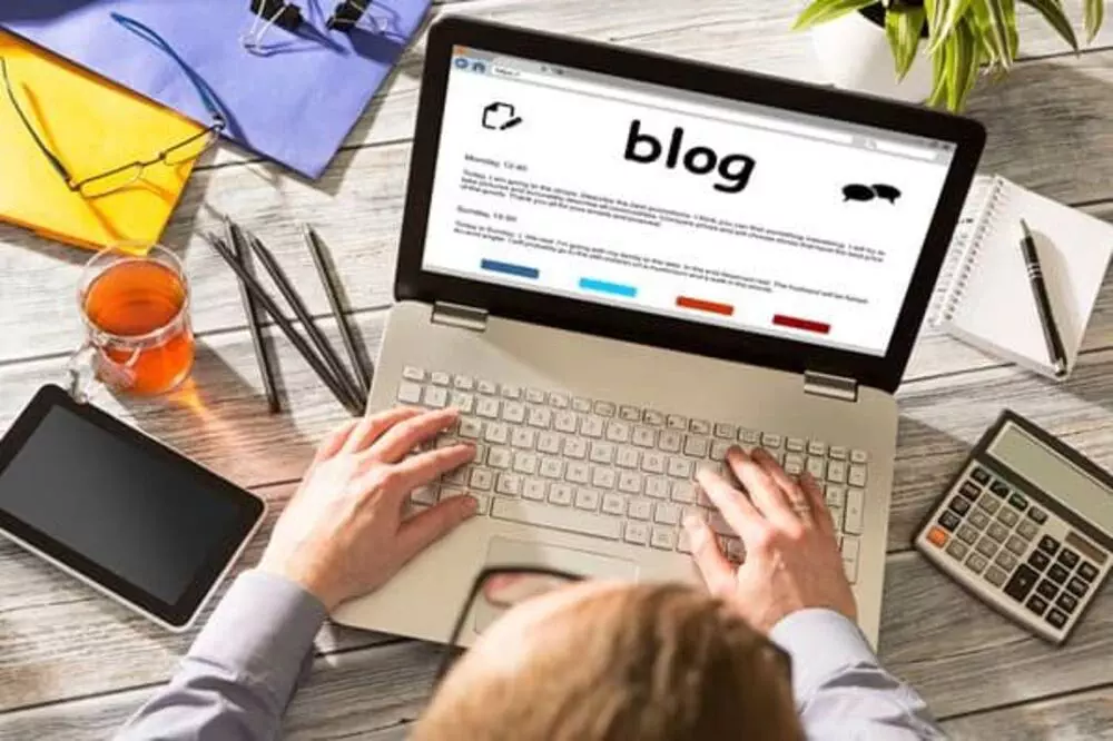 Person typing on a laptop with a blog on the screen, surrounded by a tablet, smartphone, calculator, and a cup of tea.