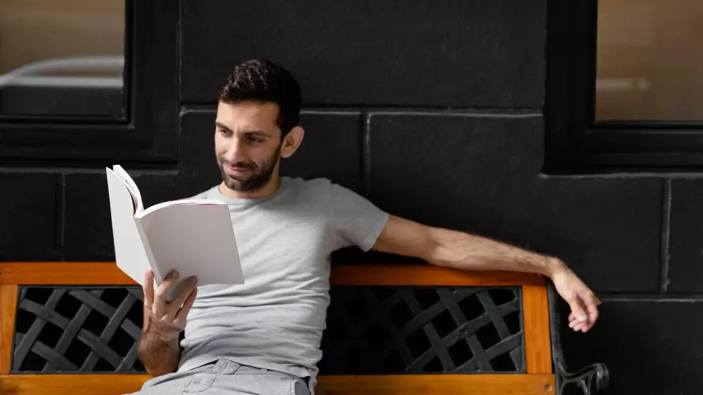 Man relaxing on a bench while reading a book in a peaceful setting against a dark wall.